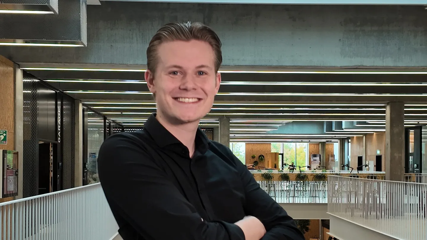 Smilende studerende i sort skjorte med armene over kors i en lys universitetsbygning